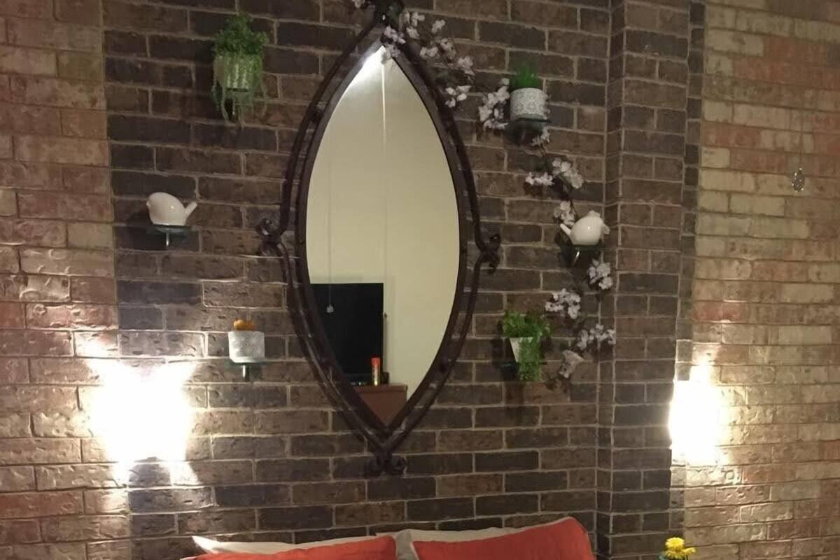 Un dormitorio con pared de ladrillo, un espejo grande, una cama con ropa de cama roja y beige, y dos mesitas de noche con pequeñas plantas.