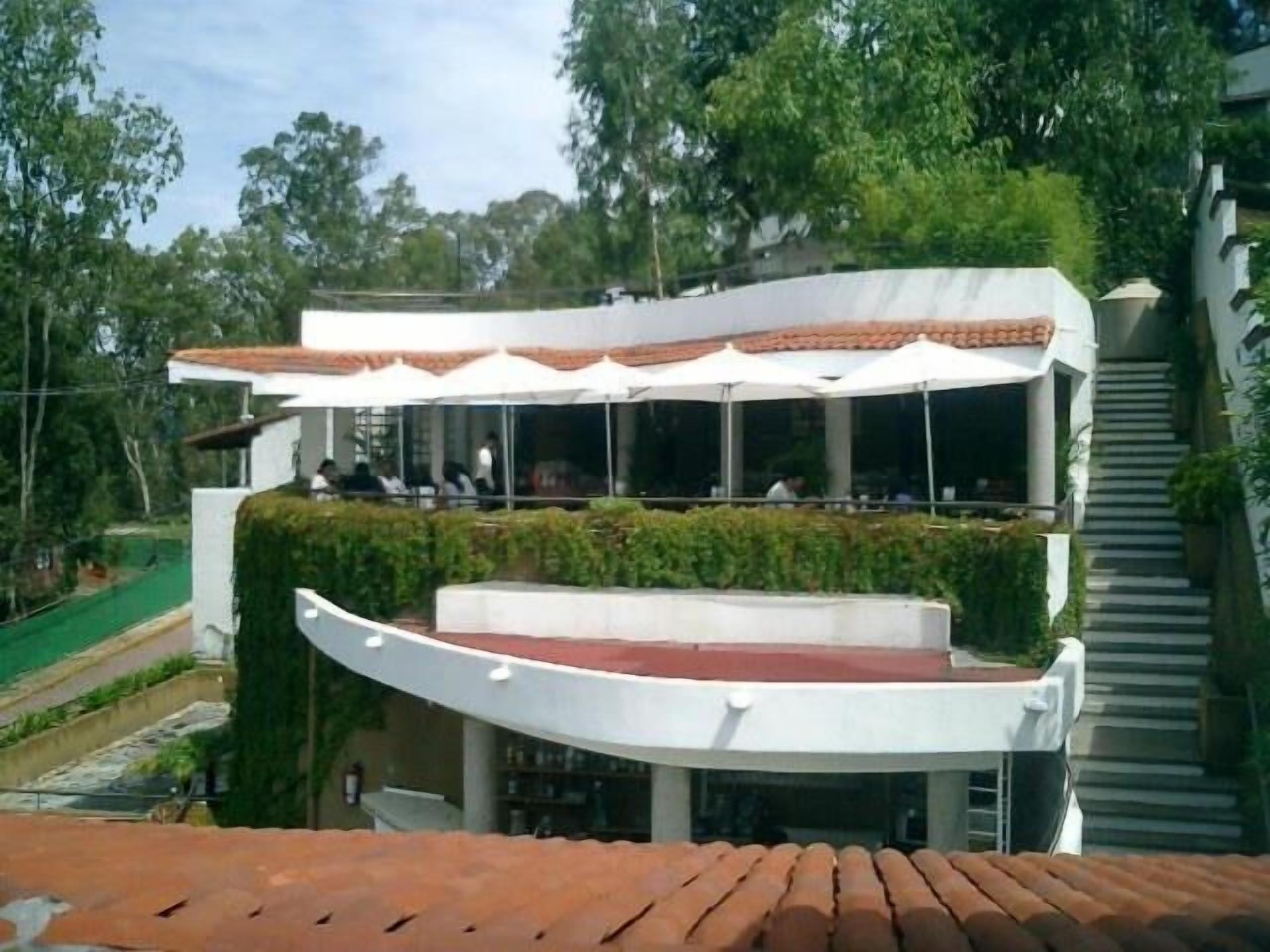 Un edificio moderno con techo curvo, ventanas amplias y mesas al aire libre bajo sombrillas blancas.