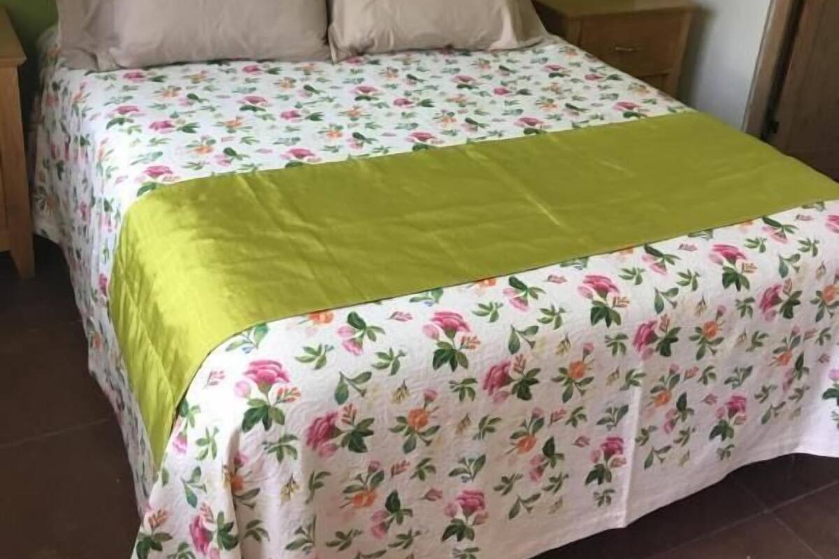 Una cama con ropa de cama floral y dos almohadas, una mesita de noche y una lámpara de pared.