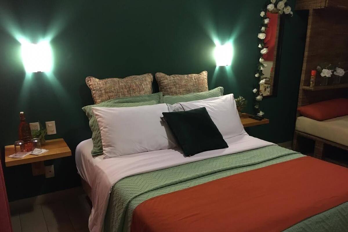 Una cama bien hecha con decoración verde y blanca, una mesita de noche de madera con una botella y vasos, y una lámpara fijada a la pared.