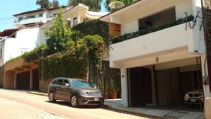 A street view with a parked SUV, a white building with a garage, and a balcony with greenery. - Casa Cielo (Valle de Bravo)