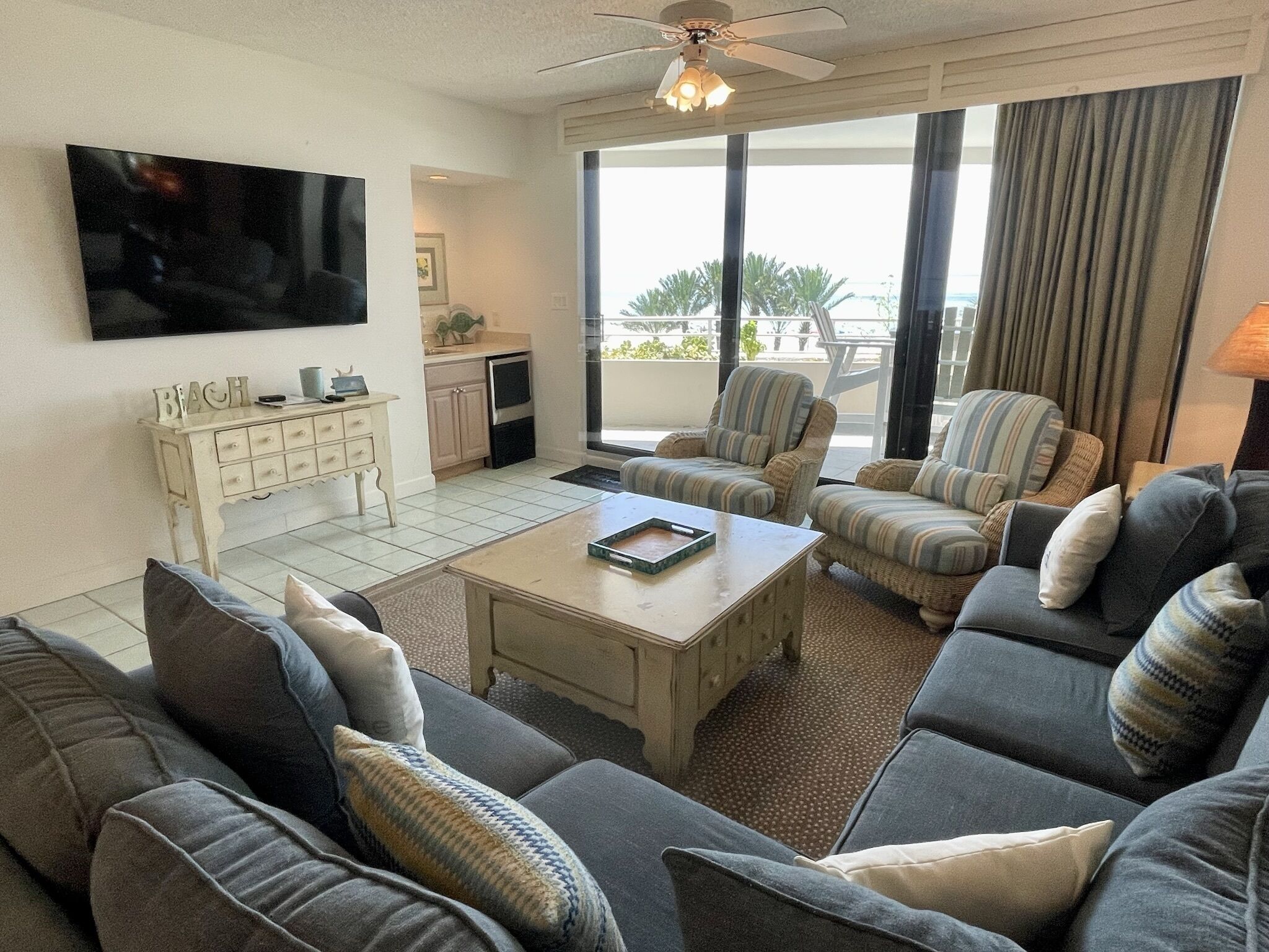 A living room with a sofa set, a coffee table, a TV mounted on the wall, and a balcony with curtains.