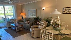 A living room with a sofa, coffee table, and a dining table with chairs. There is a lamp, a painting on the wall, and a ceiling fan.