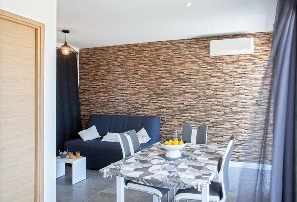 Un salon moderne avec un canapé bleu foncé, une table à manger recouverte d’une nappe à motifs et un mur en pierre.