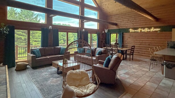A spacious room with large windows, a hanging chair, a sofa, and a dining area. - Cozy Log Cabin - Nature Escape - 5 acres (Iola)