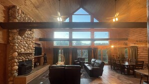 A spacious log cabin interior with a stone fireplace, leather sofas, a dining area, and large windows offering natural light and views. - Cozy Log Cabin - Nature Escape - 5 acres (Iola)