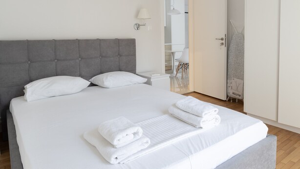 A neatly made bed with white linens and towels, a grey upholstered headboard, and a white wall with a light fixture. - Acropolis Urban Life (Athens)