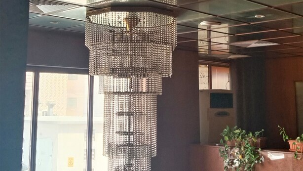 A modern interior with a large, multi-tiered chandelier hanging from the ceiling, large windows allowing natural light, and a potted plant on a wooden table. - Al Commodore Hotel (Manama)