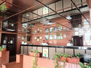 A hotel reception area with a reception desk, framed pictures on the wall, and potted plants. - Al Commodore Hotel (Manama)