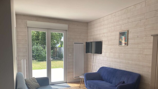 Ein modernes Wohnzimmer mit einem blauen Sofa, einem an der Wand befestigten Fernseher und einer Glastür, die in einen Garten führt.