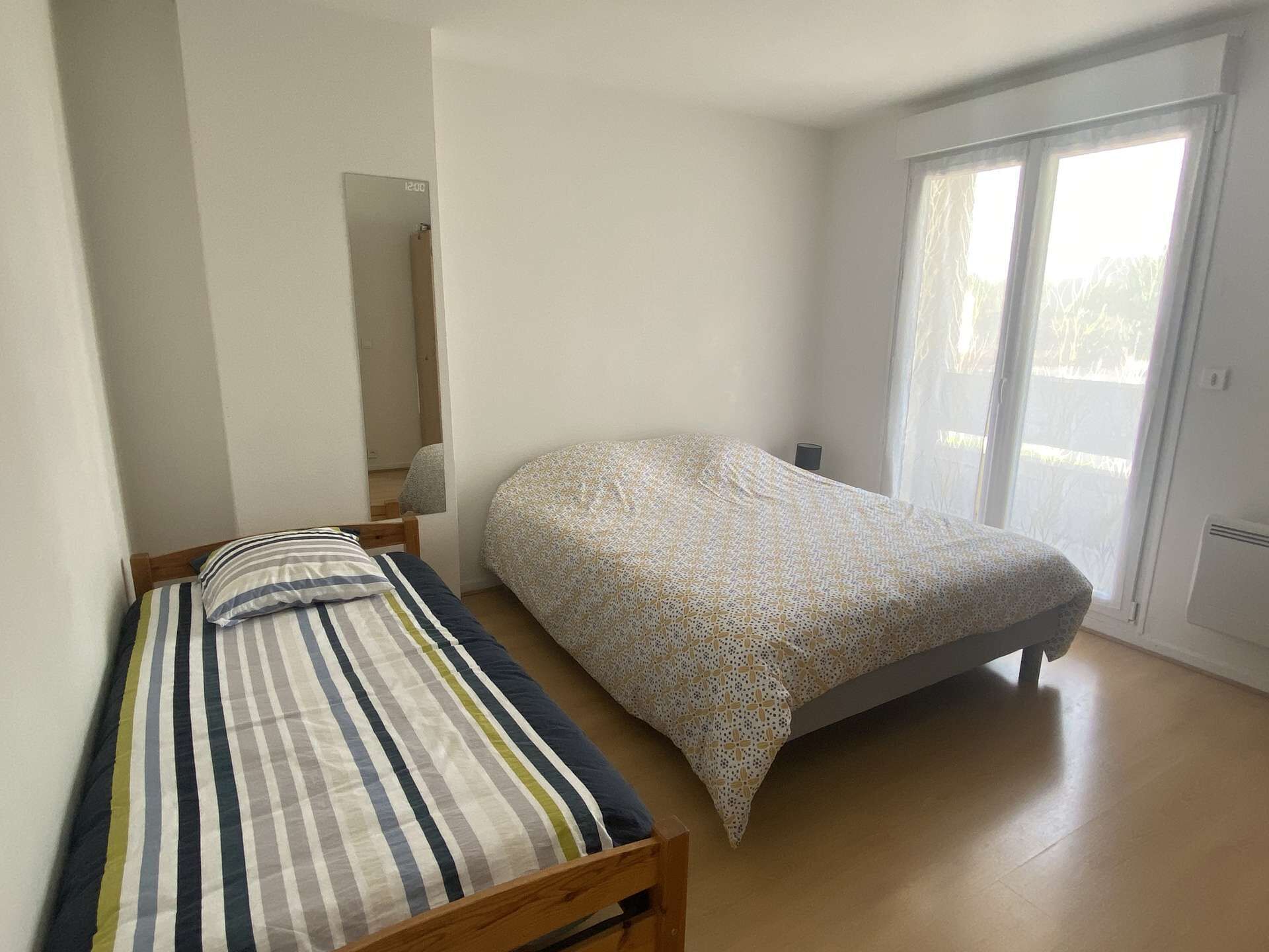 Een kleine eenpersoonskamer met een houten hoofdbord, een bed met een patroon erop, en een groot raam met uitzicht op bomen.