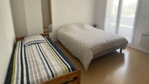 Ein kleines Zimmer mit einem Einzelbett, einem Holz-Kopfende, einer gemusterten Tagesdecke und einem großen Fenster mit Blick auf Bäume.