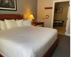 A large bed with white linens, a bedside table with a lamp, a small chair, and a door leading to another room.