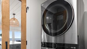 Een moderne wasruimte met twee roestvrijstalen voorladers en drogers, een houten deur en een uitzicht op een keuken met een hanglamp.