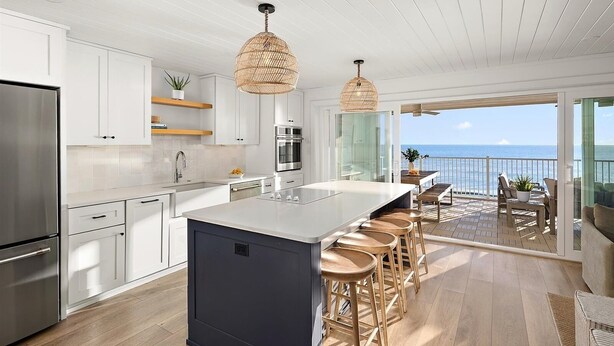 Een moderne keuken met witte kasten, een centraal eiland met krukken, een roestvrijstalen koelkast en uitzicht op de oceaan door een glazen deur.