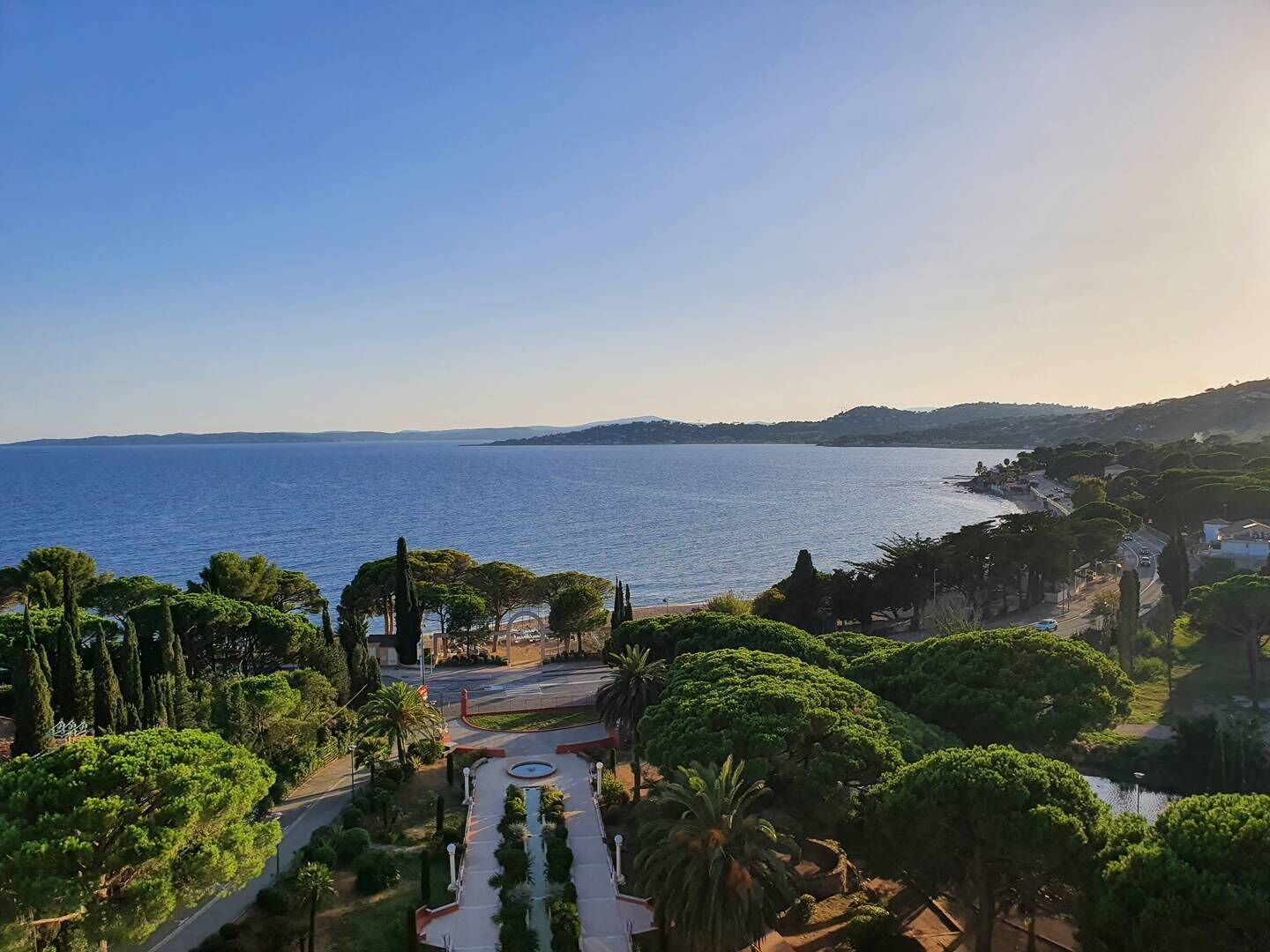 Un paysage côtier offrant une vue dégagée sur la mer, une route menant à une plage, ainsi qu’une variété d’arbres et de végétation.