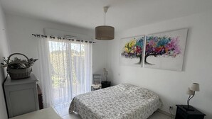 Un dormitorio con cama, escritorio, silla, lámpara, ventana con cortinas y un cuadro en la pared.