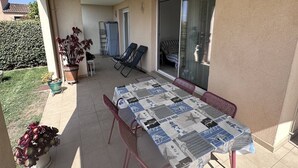 Un patio techado con una mesa dispuesta para dos personas, dos sillones de descanso, una planta en maceta y un ventanal corredizo que da a un área cubierta.