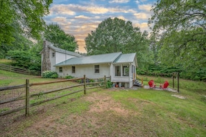 A white house with a stone chimney, surrounded by a wooden fence and a grassy yard.