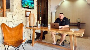 Un homme est assis à une table en bois, devant un ordinateur portable. On aperçoit un mur de pierre, un téléviseur et un ventilateur de plafond dans la pièce, qui a un sol en pierre.