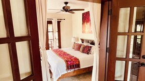 Une chambre à coucher avec un lit, un ventilateur de plafond et une vue sur l’extérieur, visible à travers une porte vitrée.