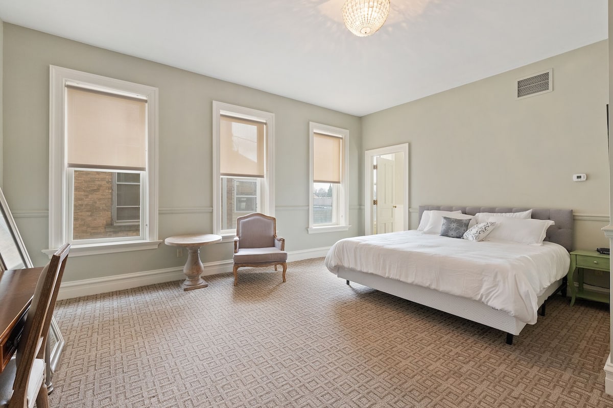 A bedroom with a large bed, a chair, a small table, and a window with blinds.