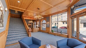 A hotel lobby with a staircase, blue armchairs, a round table, and large windows offering a view of the street.