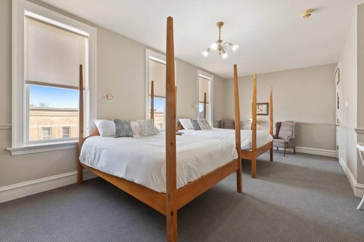 A four-poster bed in a room with large windows, a chandelier, and a chair.
