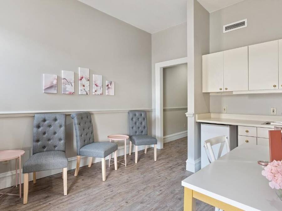 Una cocina moderna con armarios blancos, encimera blanca y un lavamanos pequeño. Hay tres sillas tapizadas con diseño capitonado, una mesa redonda rosa y una mesa de comedor blanca con un jarrón de flores.