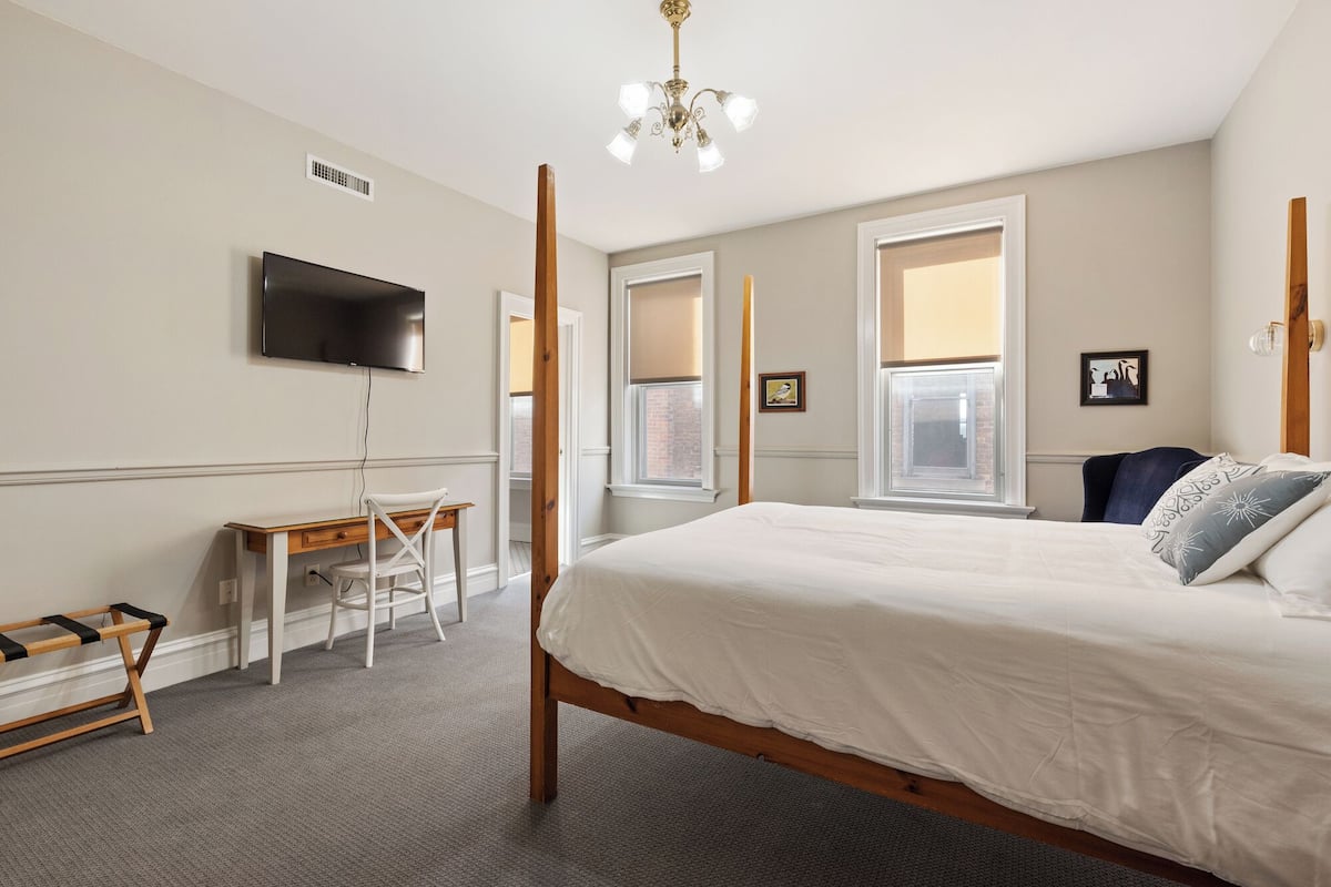 A bedroom with a bed, a desk, a chair, a TV, and a window.