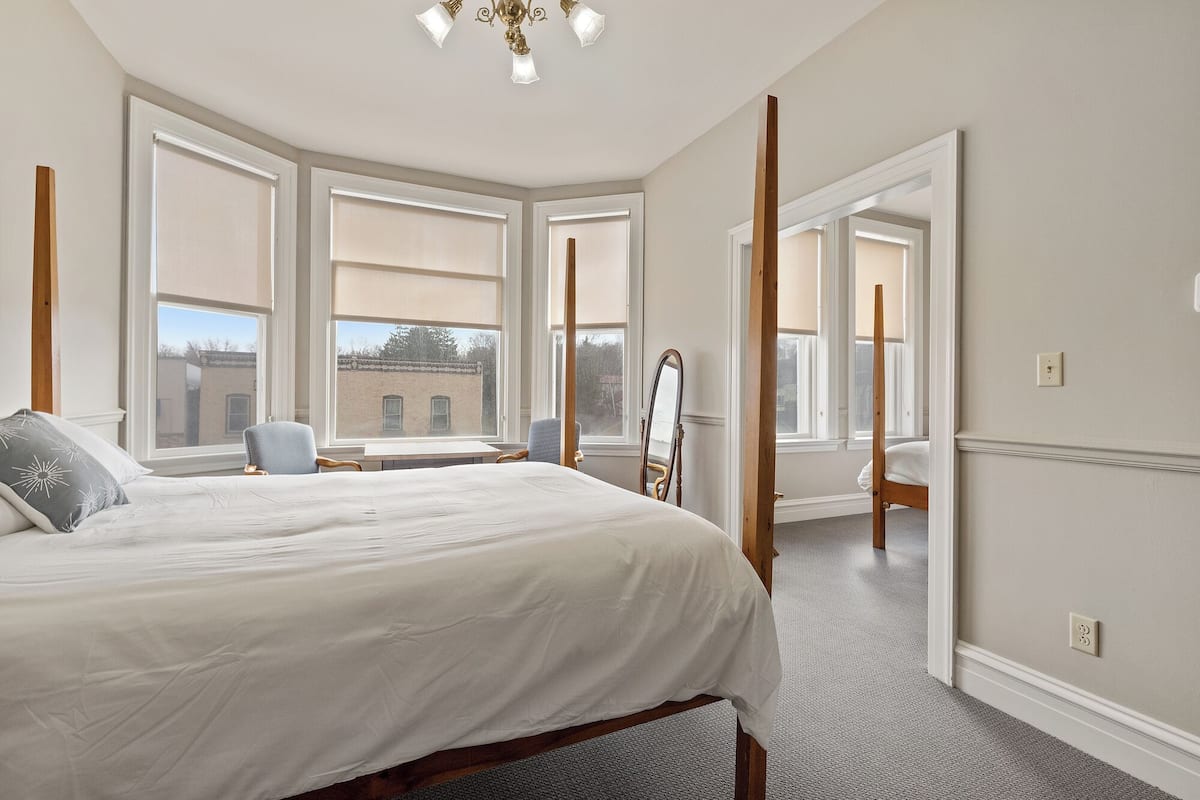 A bedroom with a large bed, a chair, a desk, and a mirror. There are multiple windows providing natural light.