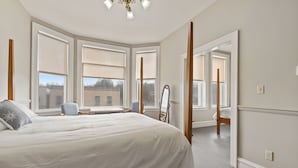 A bedroom with a large bed, a chair, a desk, and a mirror. There are multiple windows providing natural light.