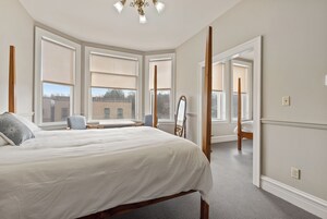 Quarto com uma cama grande, uma cadeira, uma escrivaninha e um espelho. Várias janelas proporcionam luz natural.