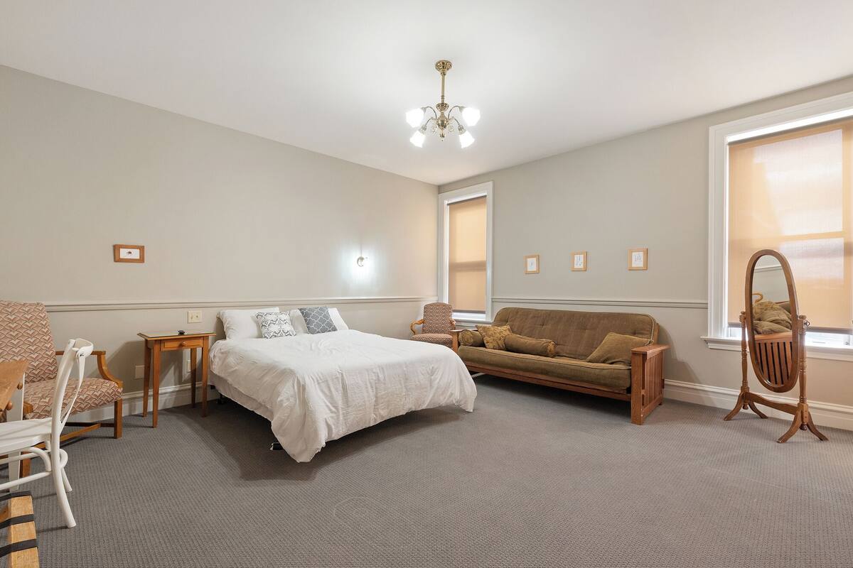 A bedroom with a bed, a sofa, a chair, a wooden side table, and a mirror.