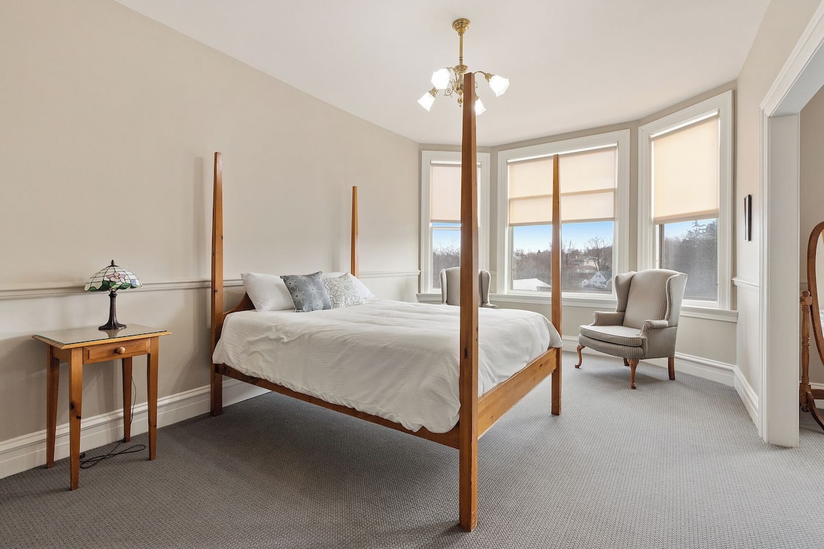 A bedroom with a four-poster bed, a bedside table with a lamp, a chair, and large windows with blinds.