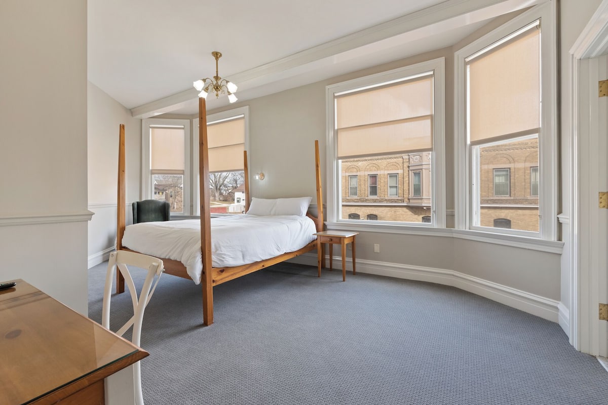 A bedroom with a four-poster bed, a desk, and large windows with city views.