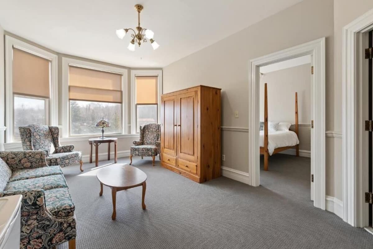 A spacious living room with a large window, a wooden cabinet, a sofa, a coffee table, and a bed in the adjacent room.