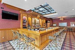 Uma área de bar com balcão de madeira, vários bancos, uma tela grande e teto de vidro.