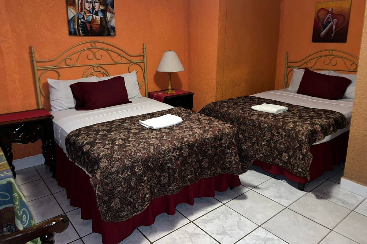 Quarto de hotel com duas camas, cada uma com colcha estampada e cortinas vermelhas. Há uma mesa de cabeceira com abajur entre as camas.