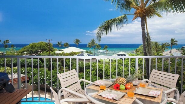 A balcony with a view of the ocean, two white chairs, a table with fruits and drinks, and a pool in the background.