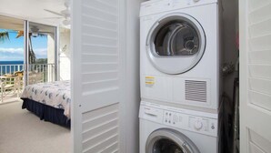 A laundry room with stacked washing machines, a bed with a blue and white floral bedspread, and a balcony with a view of the ocean.