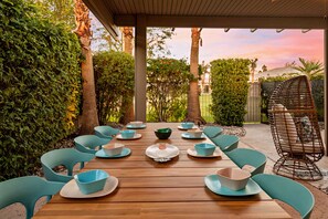 An outdoor dining area with a wooden table set for six, surrounded by teal chairs, and a wicker chair.