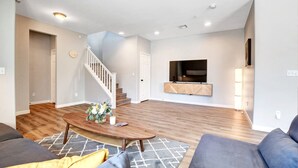 A modern living room with a staircase, wooden flooring, a flat-screen TV mounted on the wall, and a coffee table with decorative items.