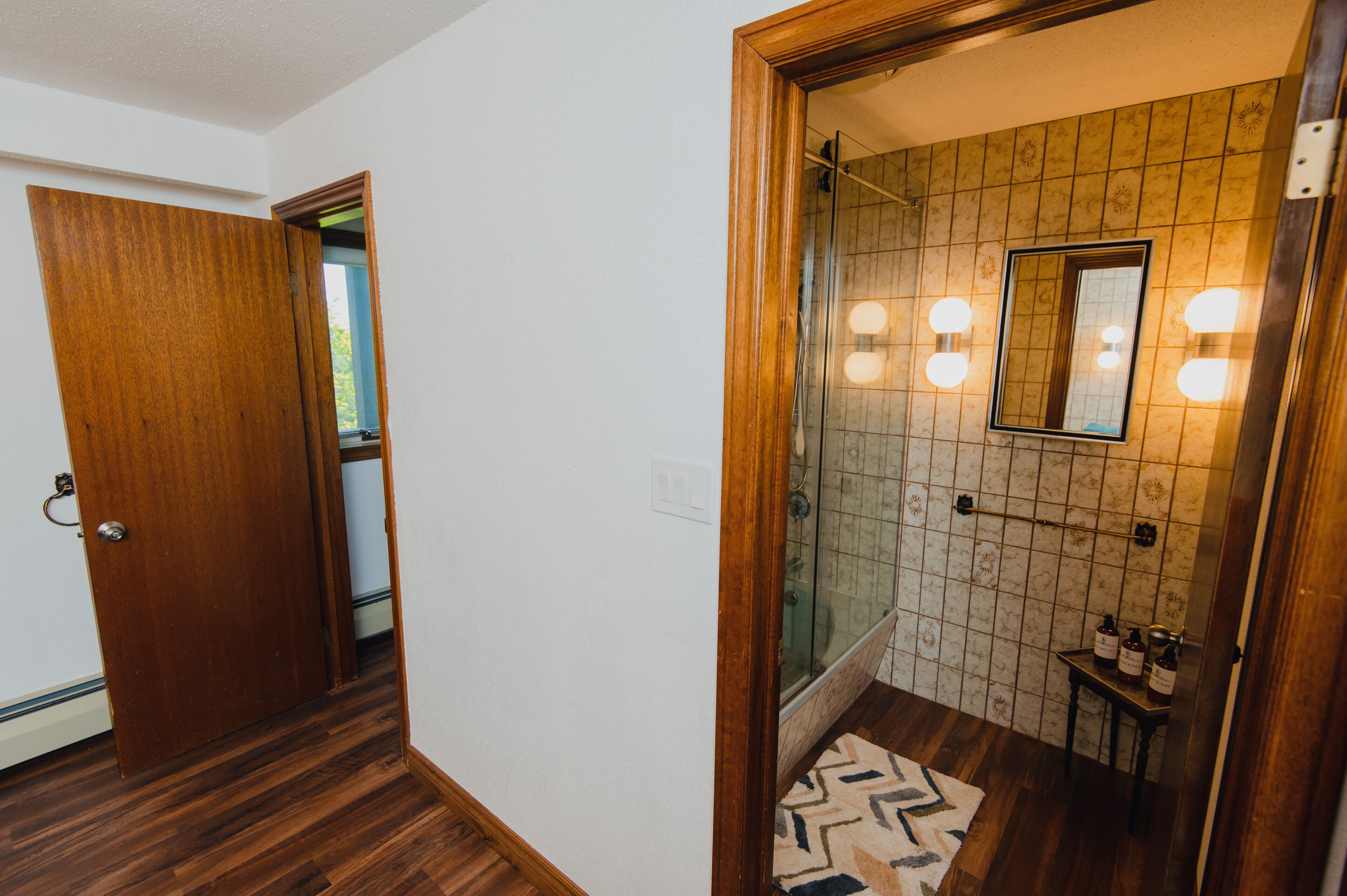 A bathroom with a glass shower enclosure, a wooden door, and a tiled wall with a mirror and lights.