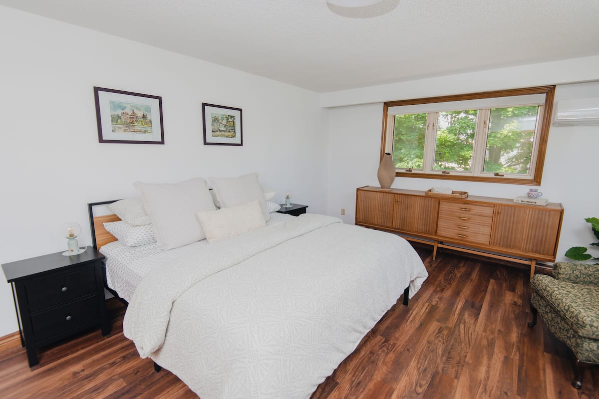 A bedroom with a large bed, a nightstand, a dresser, and a window with a view of trees.