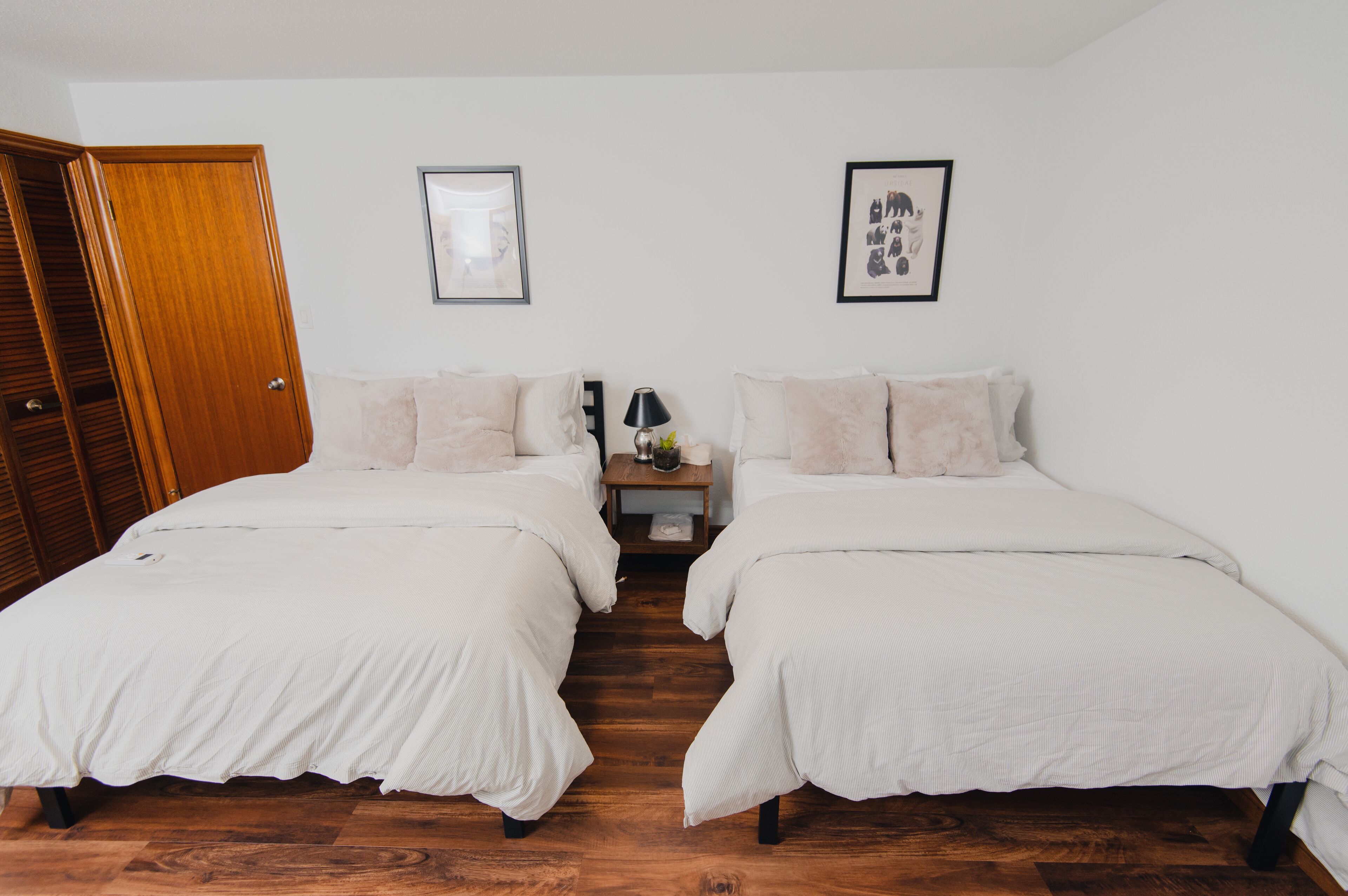 A room with two beds, wooden flooring, a nightstand with a lamp, and a door.