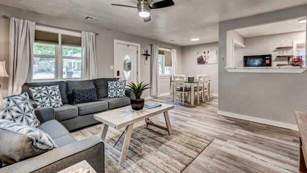 Un salon moderne avec un canapé gris, des coussins à motifs, une table basse en bois et un coin repas avec des chaises blanches.