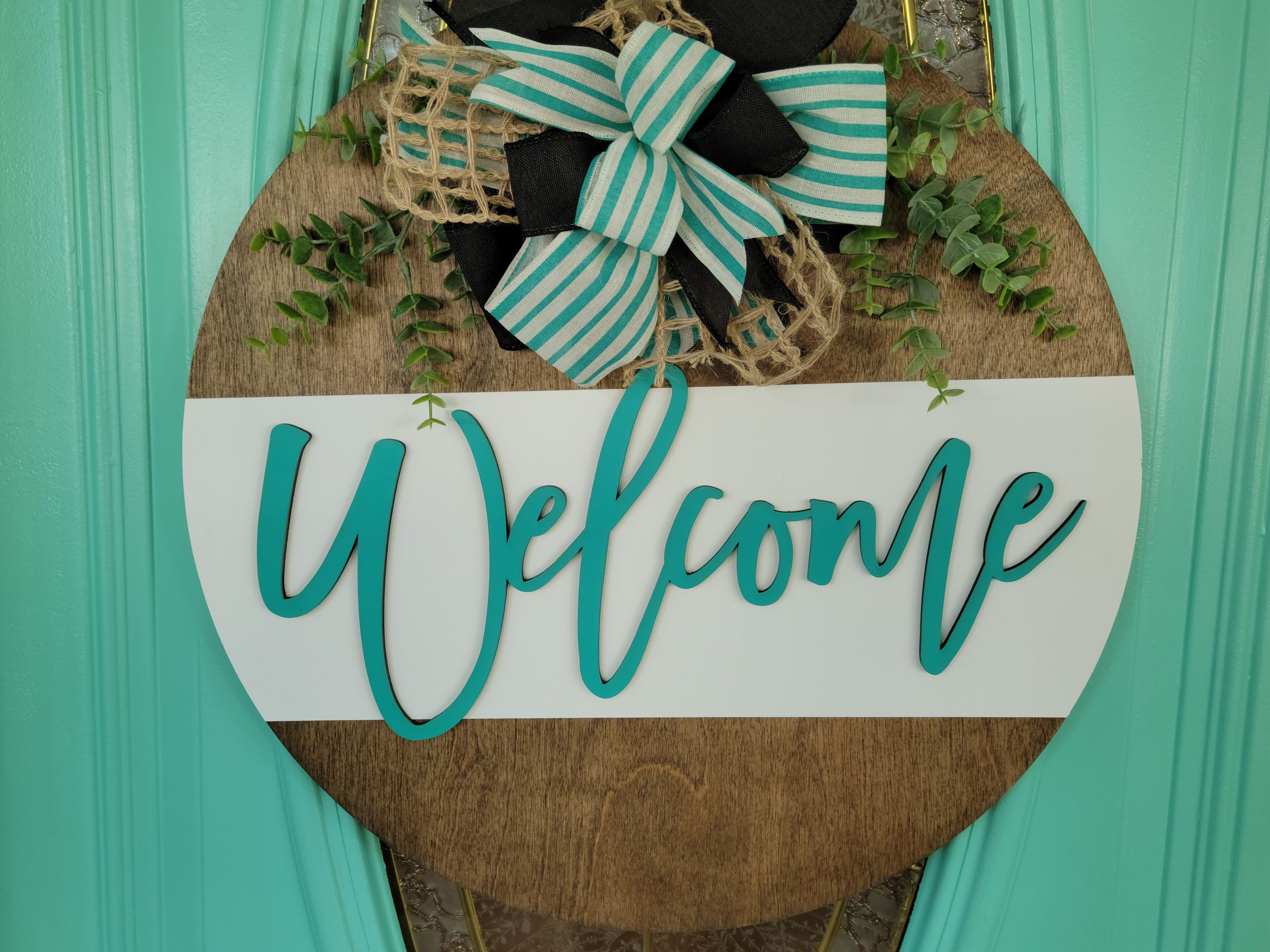 A welcome sign with a rustic wooden background and a white banner displaying the word "Welcome" in teal.