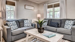 Un salon avec deux canapés gris, des coussins à motifs, une table basse en bois, une plante en pot et une citation encadrée accrochée au mur.