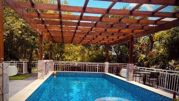 Una piscina rectangular con borde de azulejos, rodeada por una cerca blanca, bajo una pérgola de madera, con árboles al fondo.